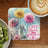 Dandelion art with inspirational text "Love grows kindness" on a coaster next to coffee and kiwi.