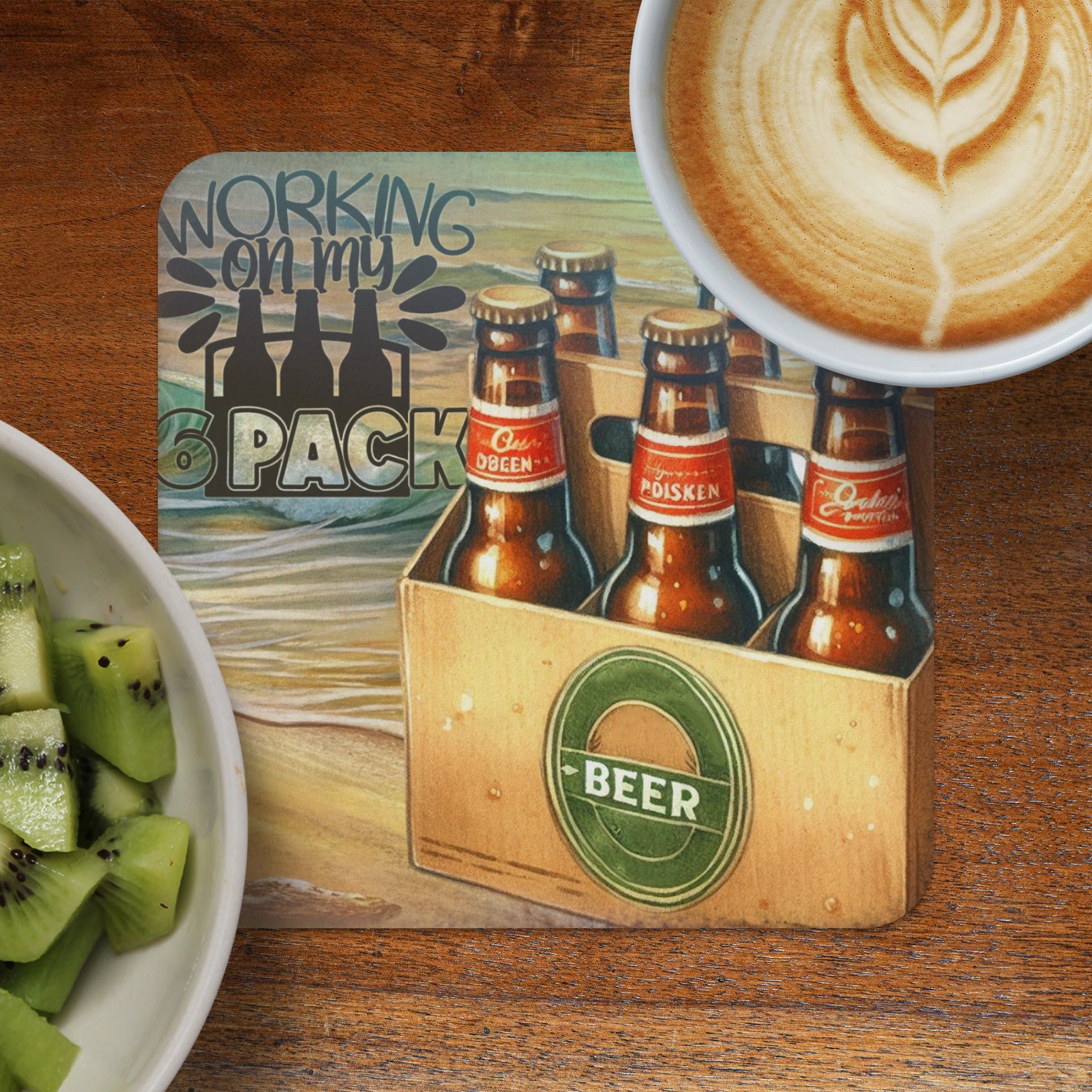 Six pack of beer bottles in a cardboard carrier on a beach-themed mat, with coffee and kiwi.