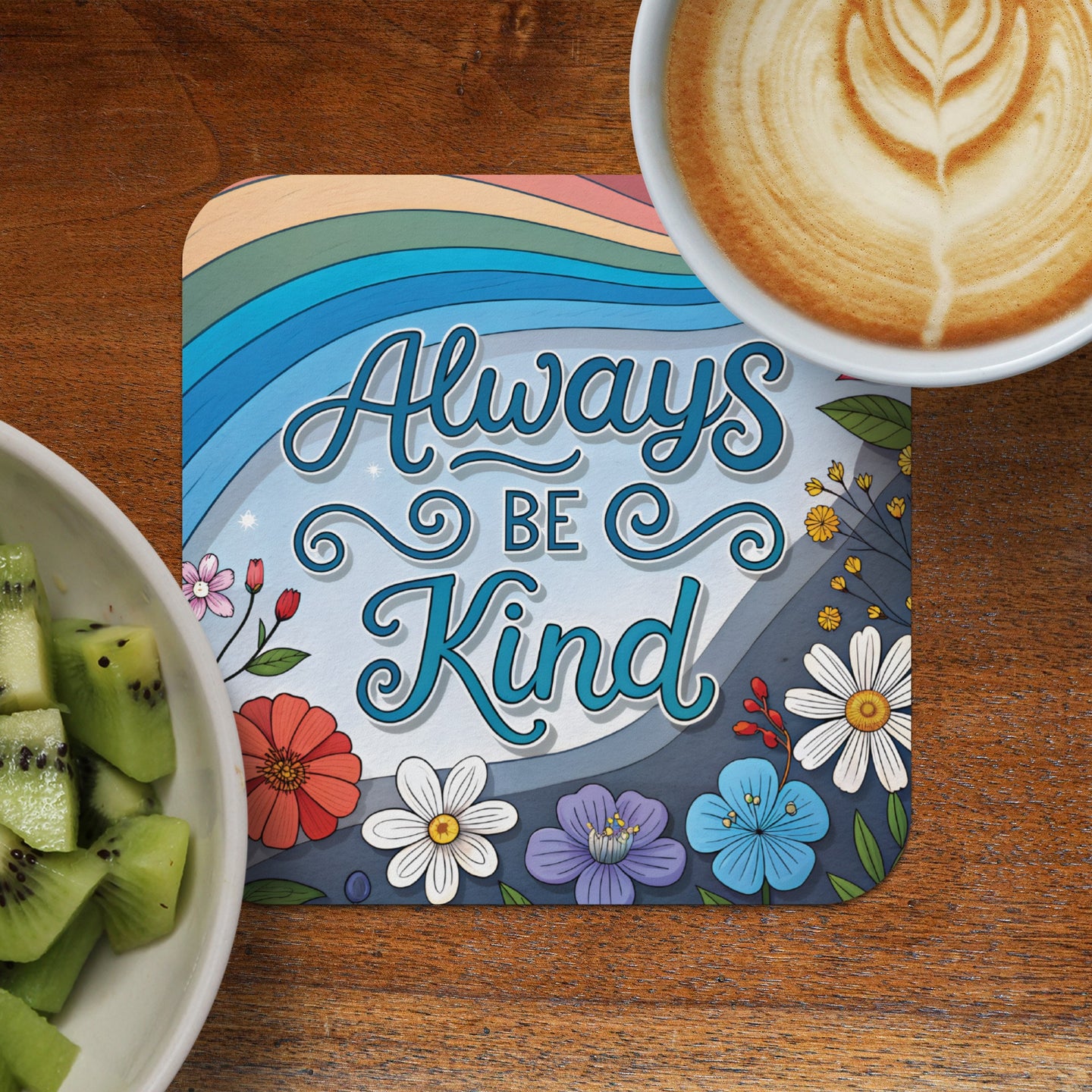 Coaster with "Always Be Kind" text and colorful flowers on wooden table with kiwi and coffee.