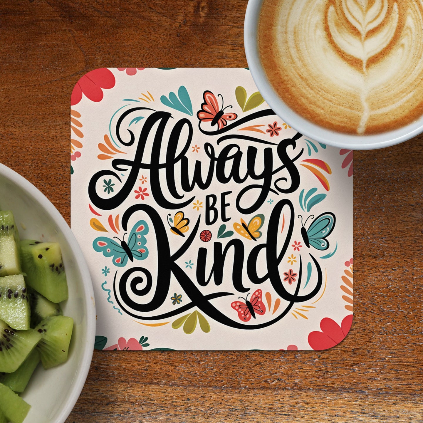 Square coaster with "Always Be Kind" text and butterflies, next to kiwi slices and coffee.