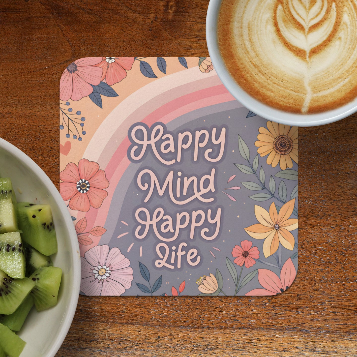 Coaster with "Happy Mind Happy Life" text and floral rainbow design, next to coffee and kiwi.