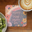 Coaster with "Happy Mind Happy Life" text and floral rainbow design, next to coffee and kiwi.