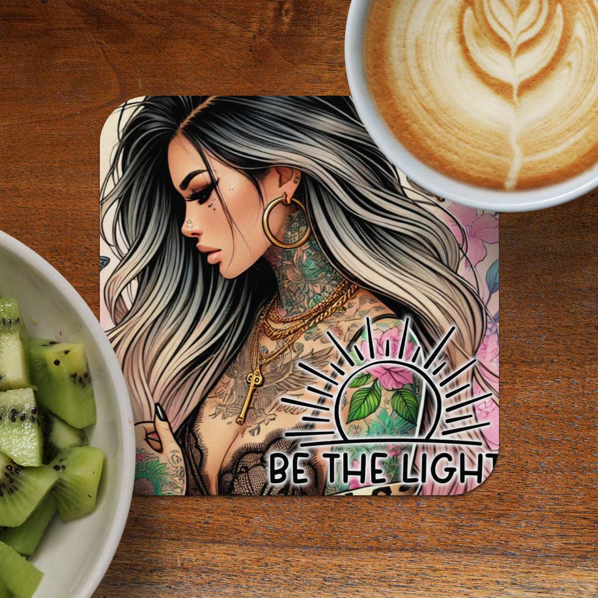Coaster with tattooed woman and "Be The Light" text, beside coffee and kiwi.