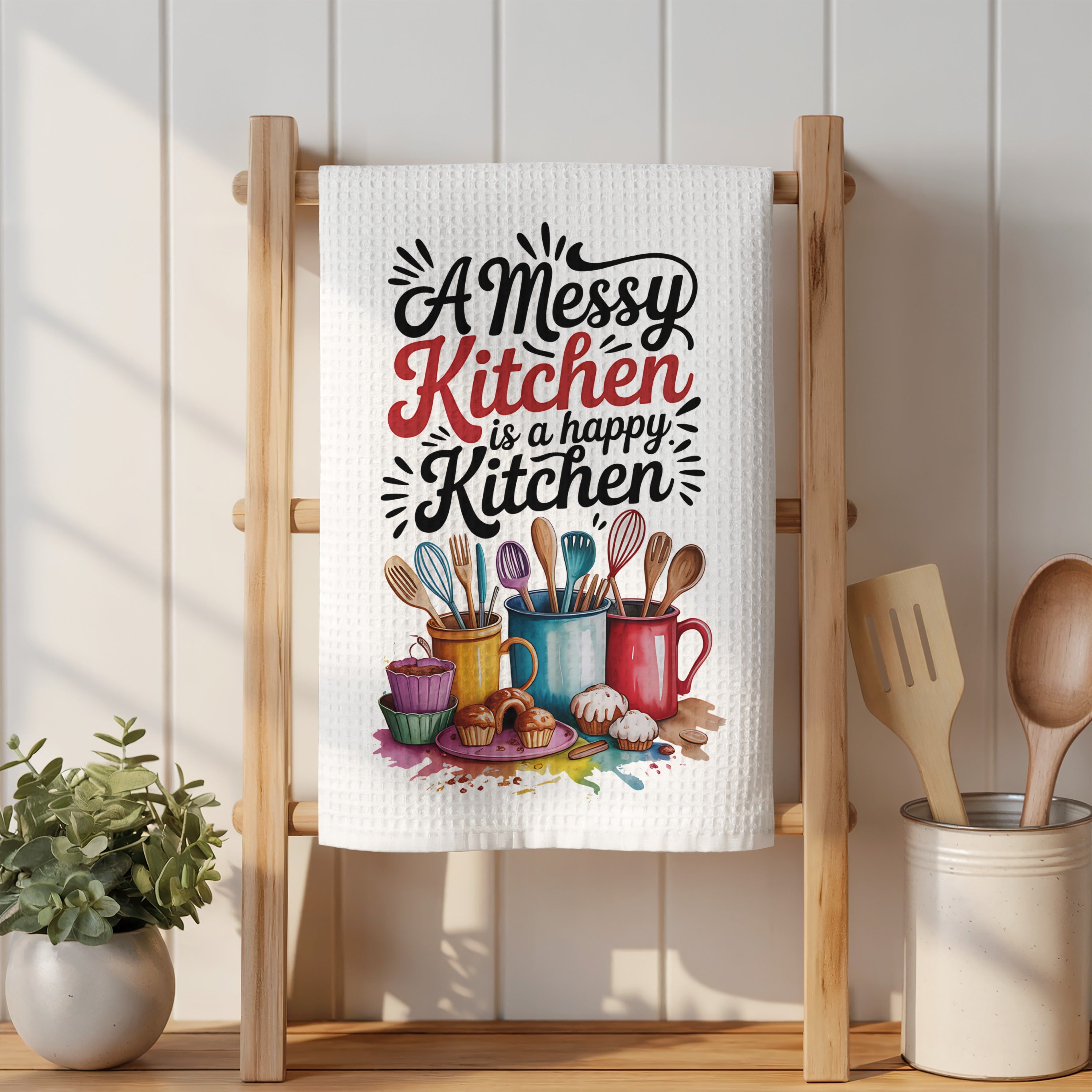 Kitchen towel with "A messy kitchen is a happy kitchen" text and colorful mugs and cupcakes.