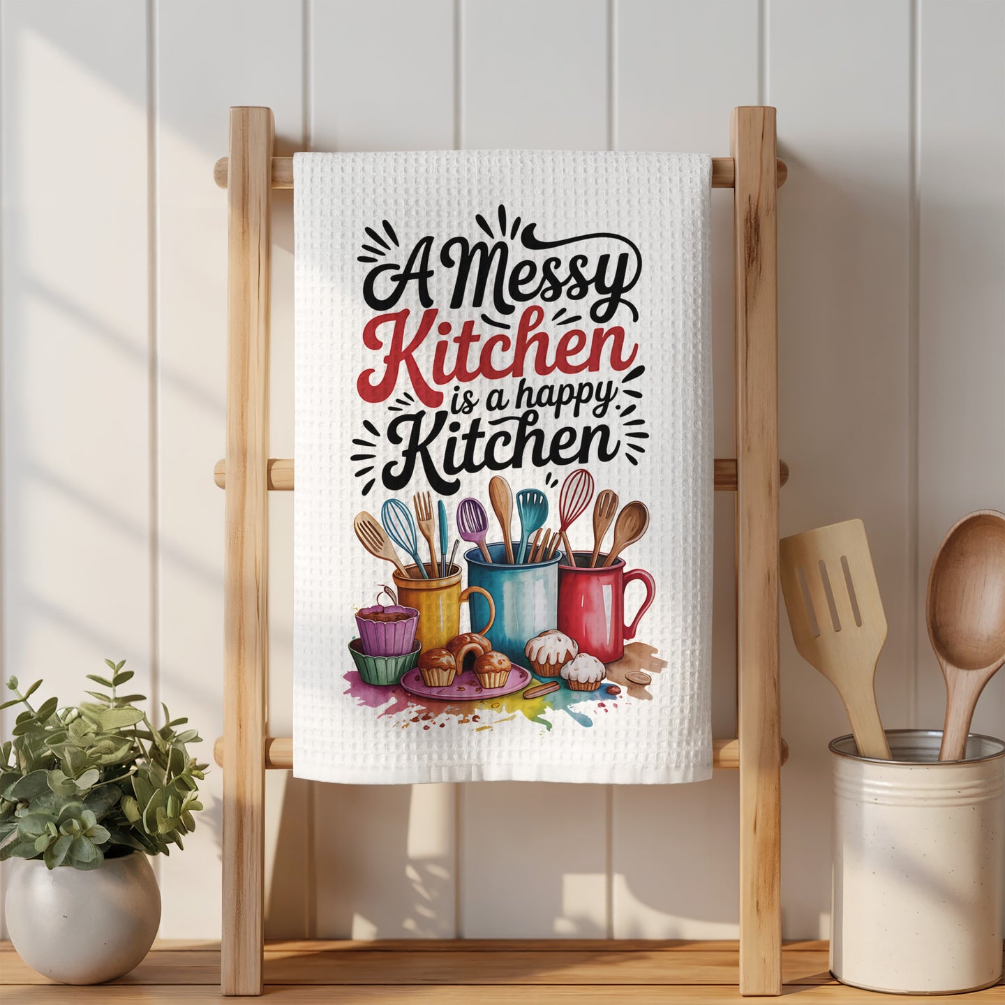 Kitchen towel with "A messy kitchen is a happy kitchen" text and colorful mugs and cupcakes.