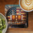 Two whiskey bottles on a barrel with an American flag background, beside fruit and coffee.