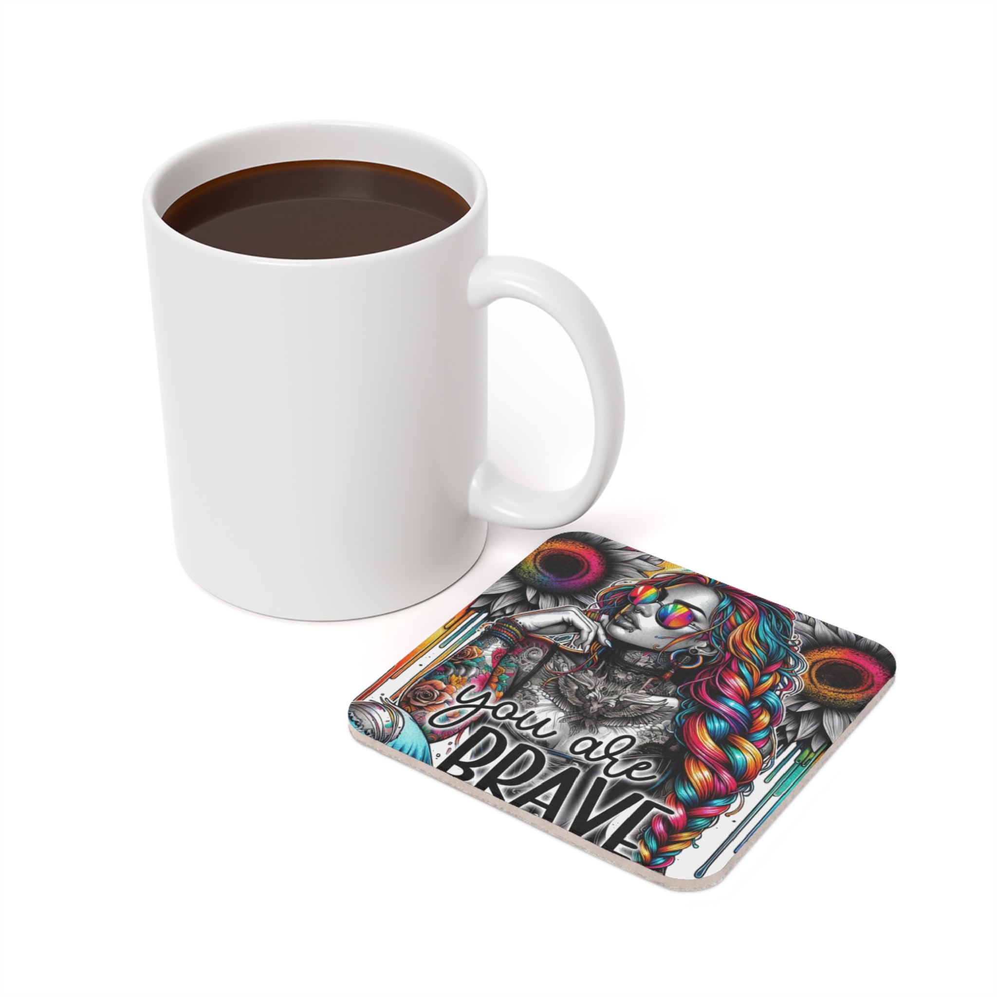White mug beside a coaster with a colorful woman and "YOU ARE BRAVE" text.