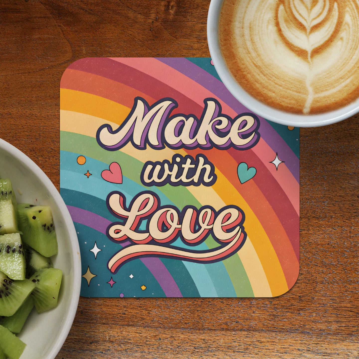 Coaster with "Make with Love" text on rainbow background next to coffee and kiwi.