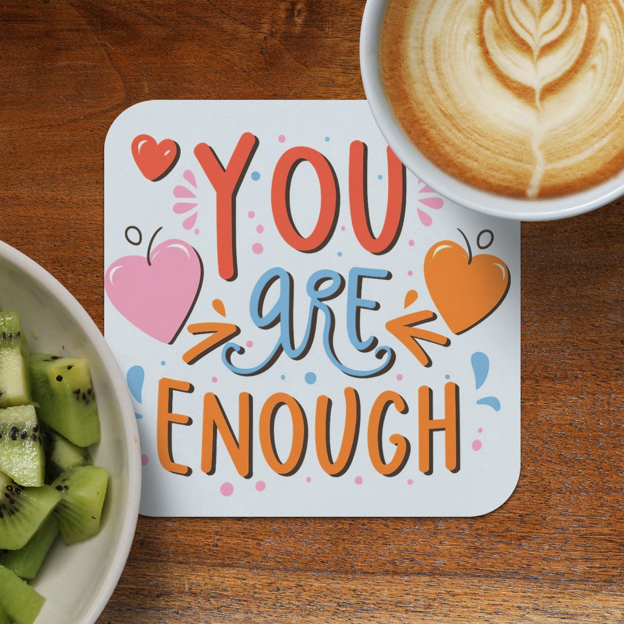 "You are enough" quote with hearts on a coaster next to a kiwi fruit bowl and coffee.