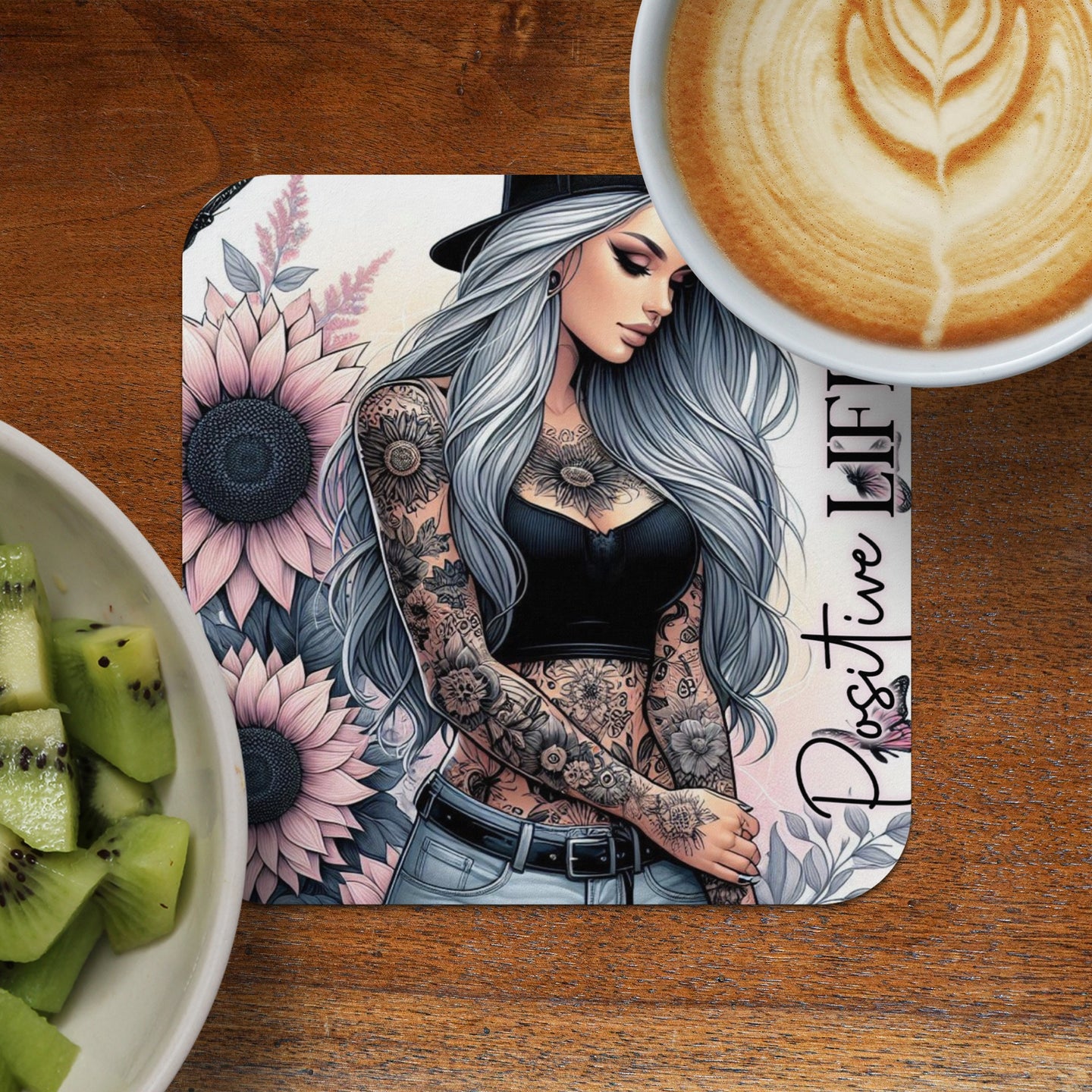Woman with tattoos and sunflowers on a coaster next to coffee and kiwi.