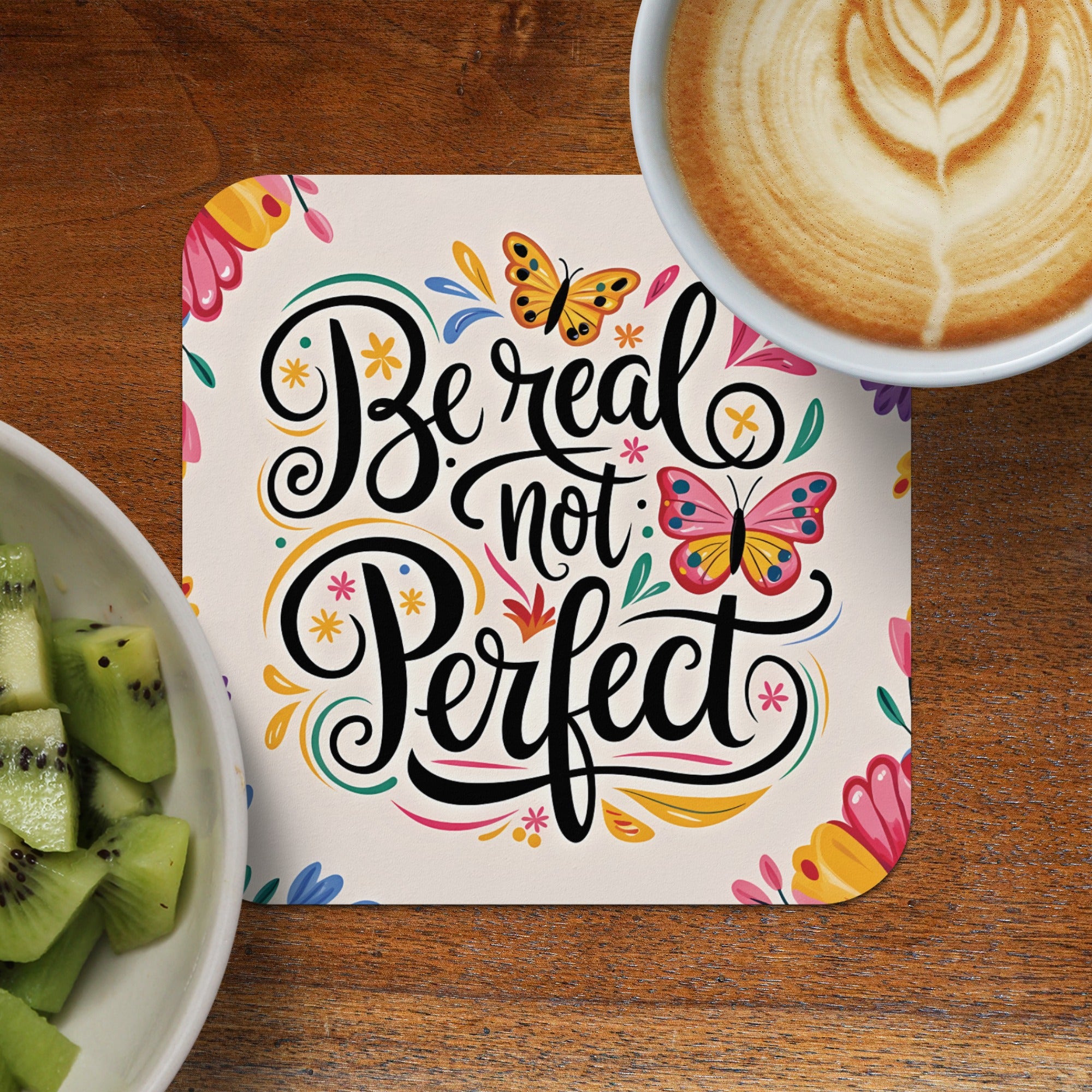 Coaster with "Be real not perfect" text and butterflies, next to kiwi and coffee.