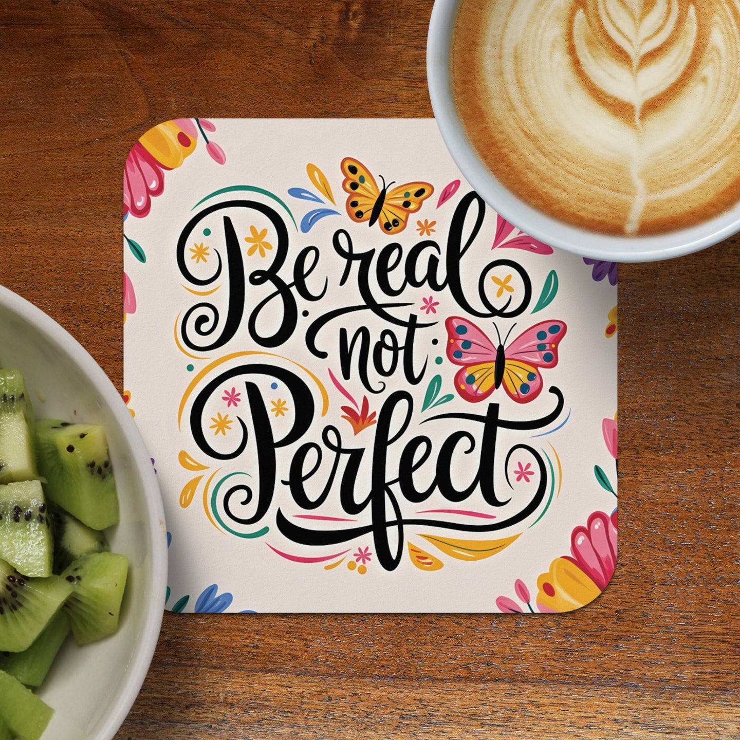 Coaster with "Be real not perfect" text and butterflies, next to kiwi and coffee.
