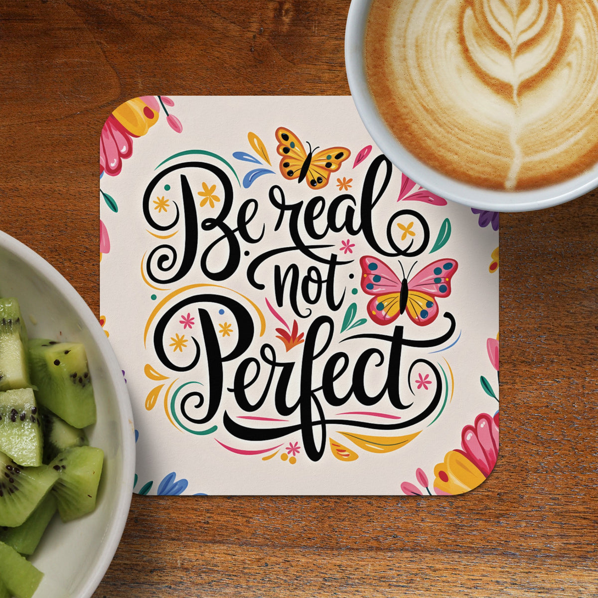 Coaster with "Be real not perfect" text and butterflies, next to kiwi and coffee.