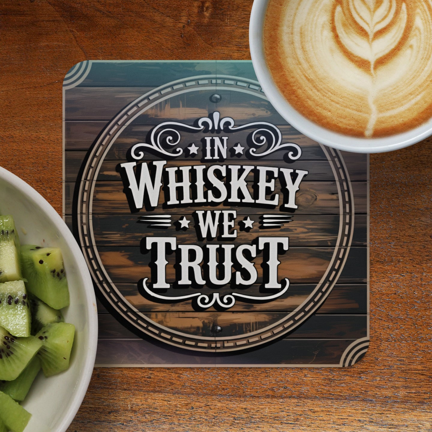 Coaster with "In Whiskey We Trust" text on wood background, next to coffee and kiwi.