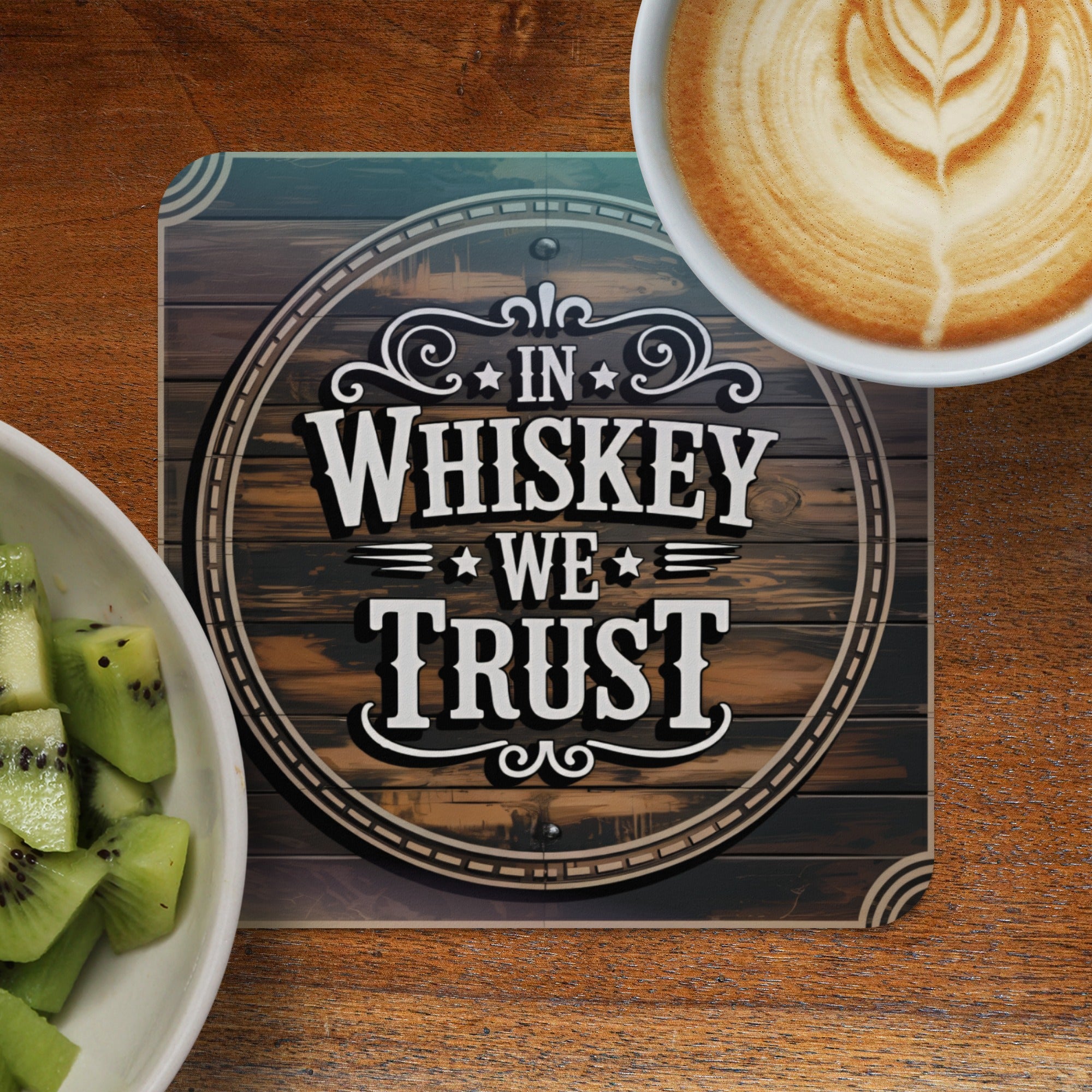 Coaster with "In Whiskey We Trust" text on wood background, next to coffee and kiwi.