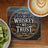 Coaster with "In Whiskey We Trust" text on wood background, next to coffee and kiwi.